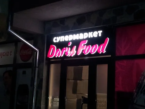 Светеща табела „Doris Food“ над входа на супермаркет, нощем